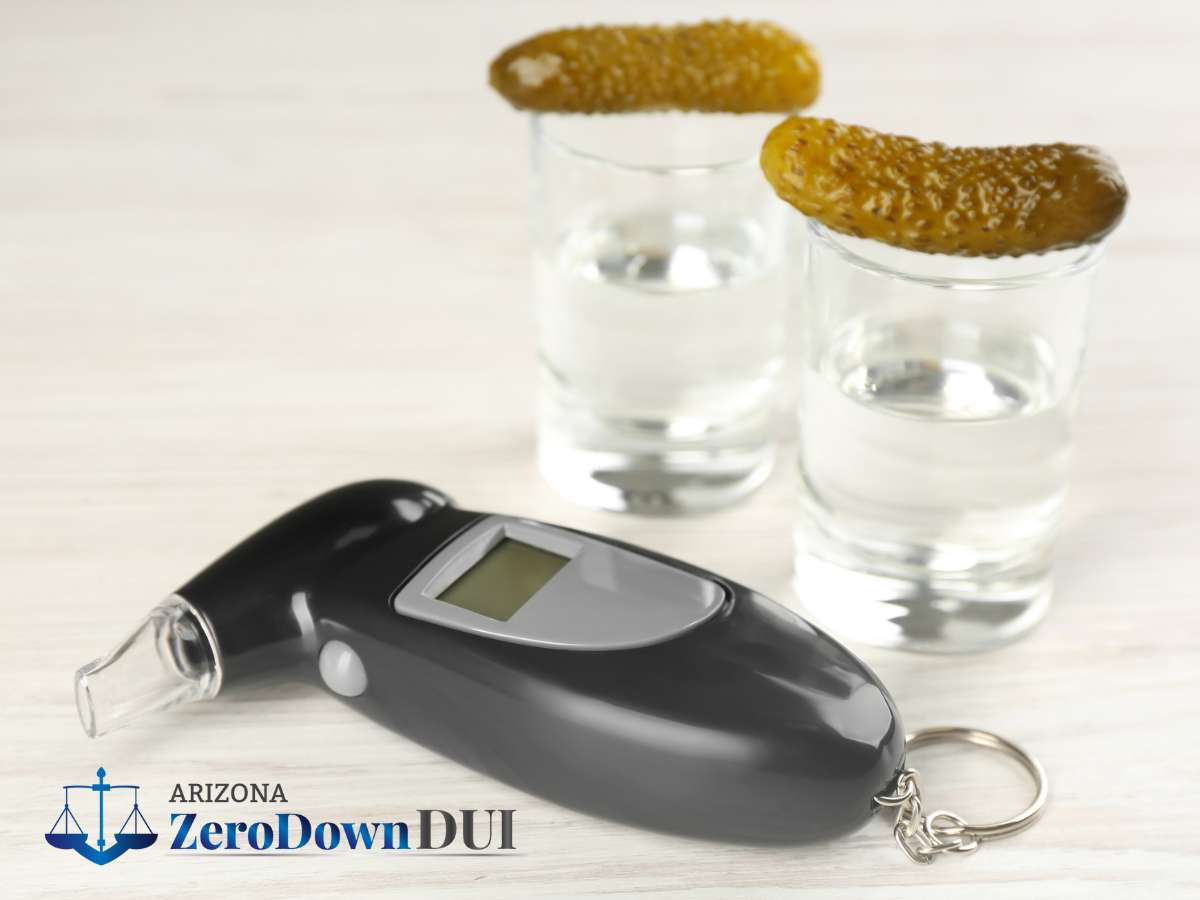 A personal breathalyzer lies in front of two shot glasses with pickles on a wooden surface.